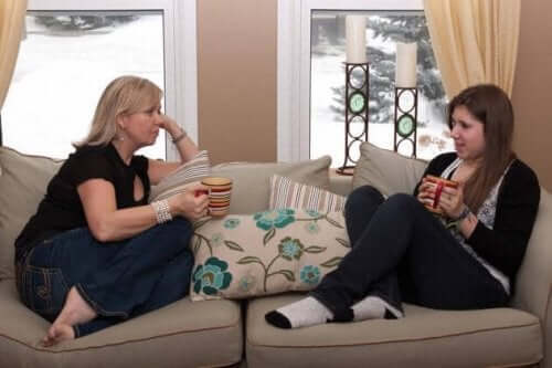 A mother and her teen daughter sitting on the couch drinking coffee and talking.