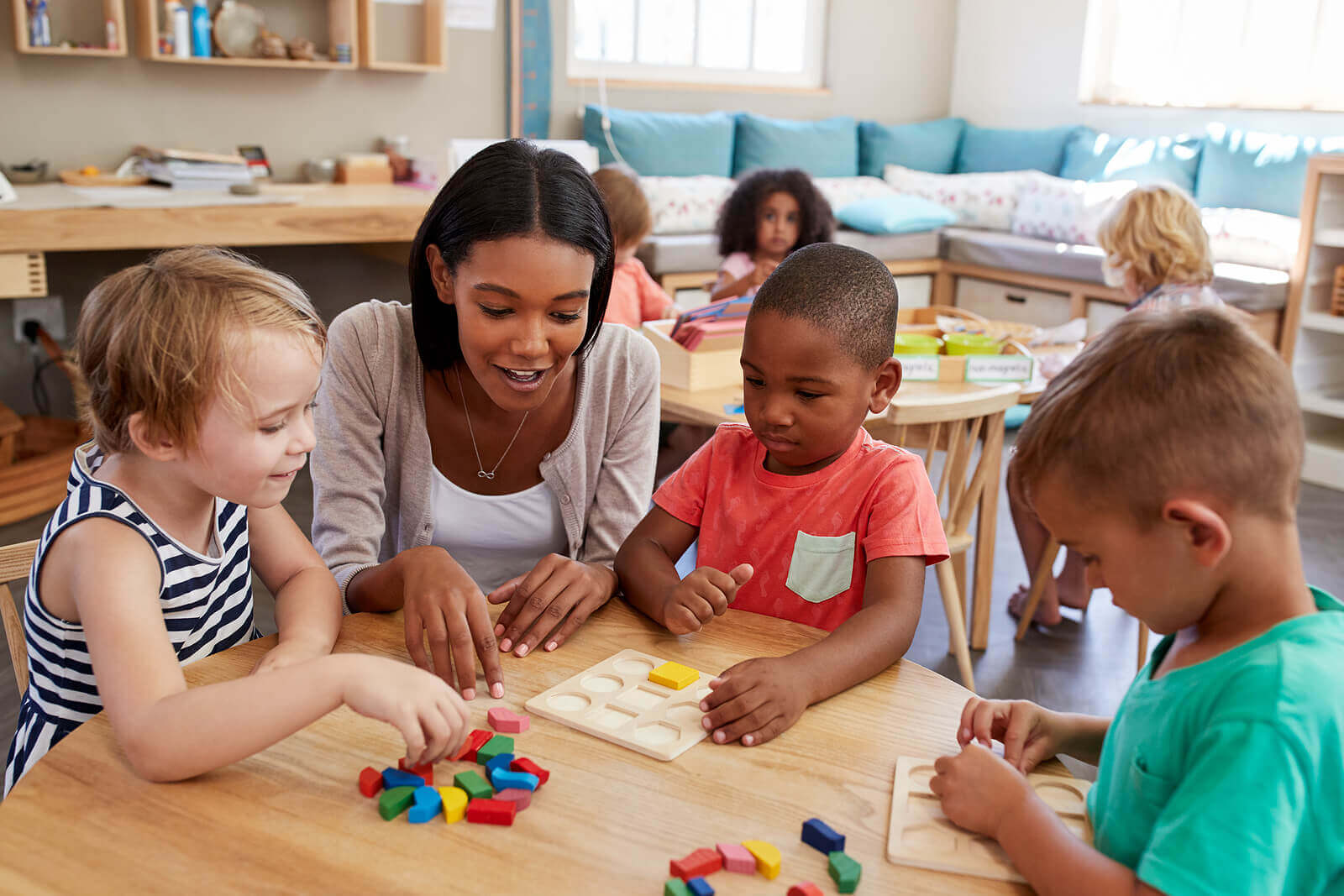 A preschool teacher sitting at a table with some students.