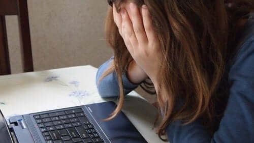A teen girl looking at a computer screen and crying into her hands.