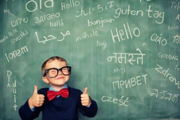 A little boy with both thumbs up standing in front of a chalkboard full of words in different languages.