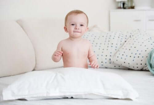 A naked baby sitting on a bed behind a pillow.