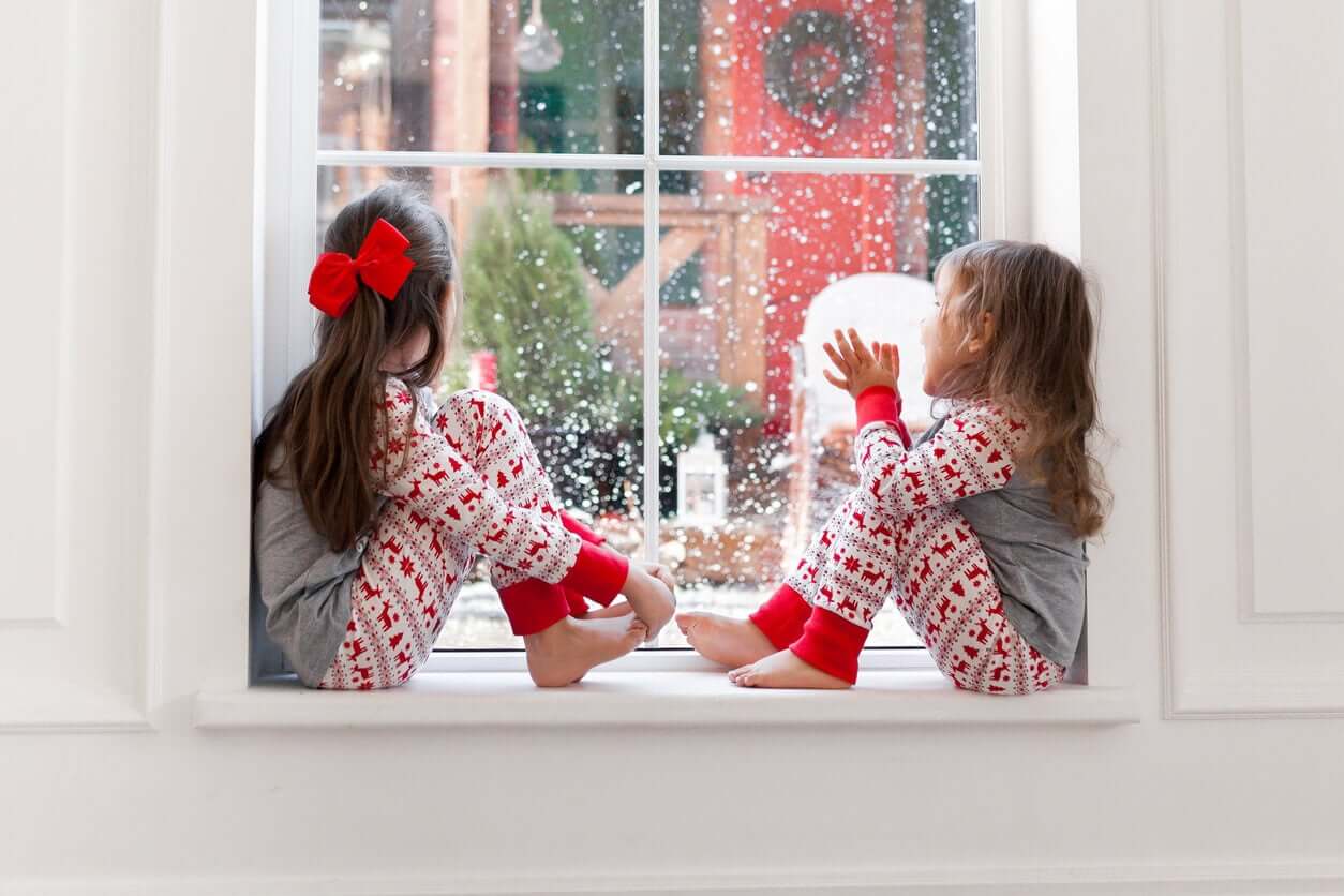 Two little girls wearing winter pajamas while sitting by a window watching the snow.
