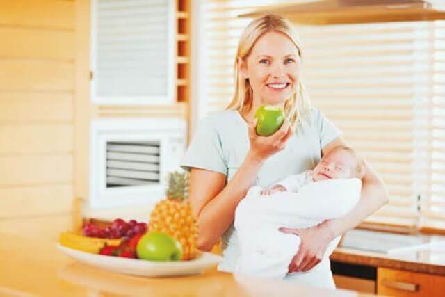 A woman holding a newborn while eating a green apple.