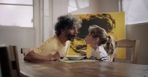 A father and daughter sucking on opposite ends of the same spaghetti noodle.