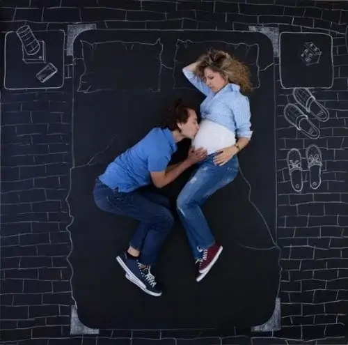 A father kissing his parther's pregnant belly as they both lie on a chalk drawing of a bed.