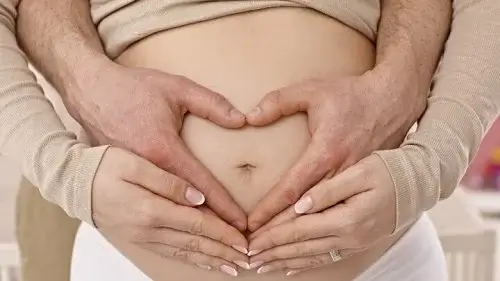 A partner hugging his partner from behind, making a heart with his hands over her pregnant belly.