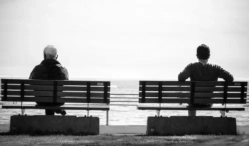 A man and his father sitting on seaparate park benches, looking out on the water.