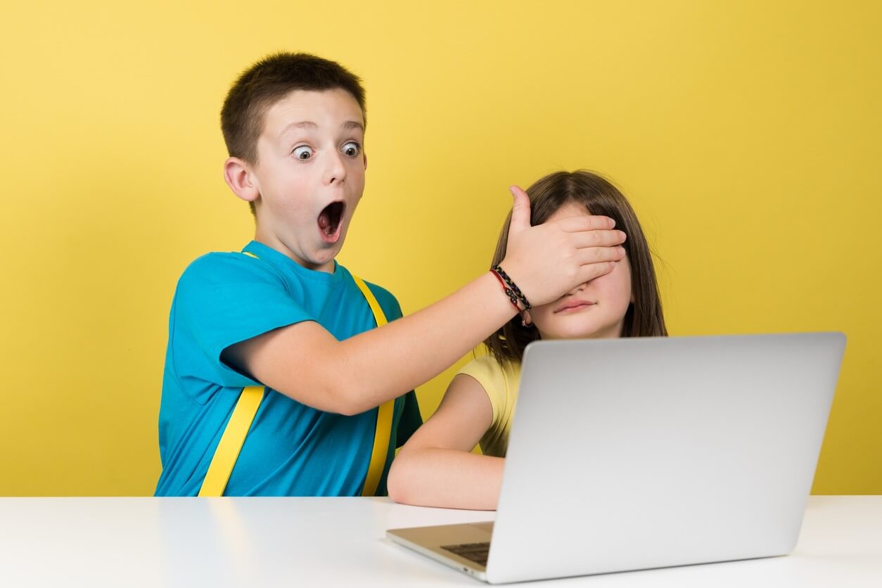 A boy covering a girl's eyes while they look at a laptop.