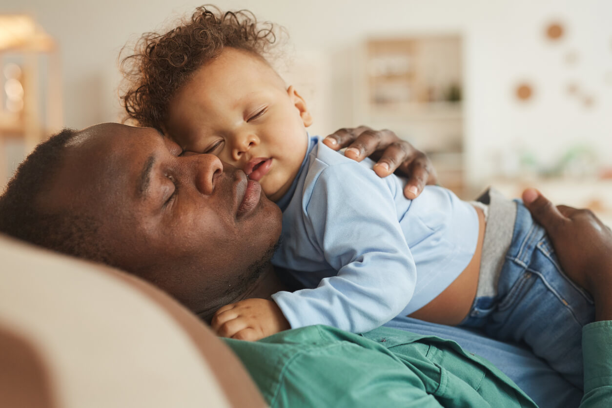 A black father taking a nap with his baby boy.