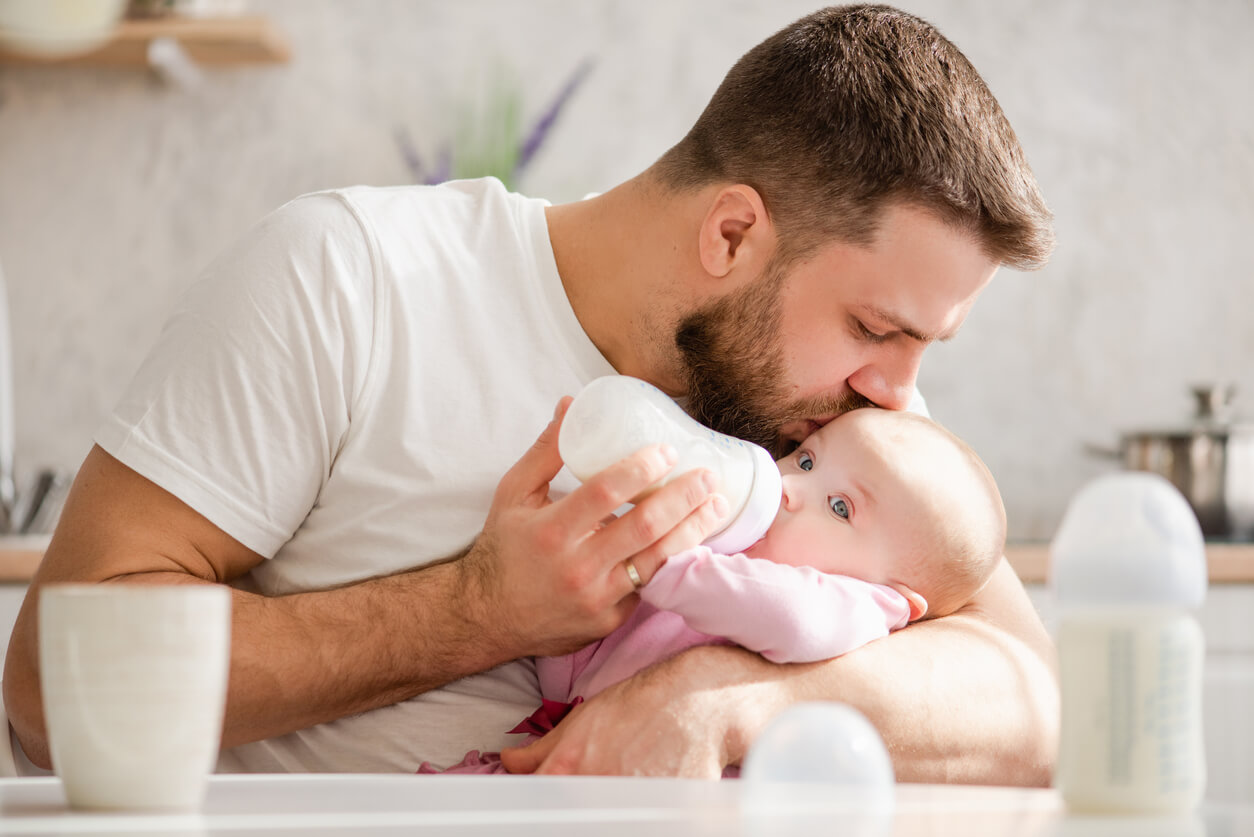 A man kissing baby's forehead while feeding her with a bottle.