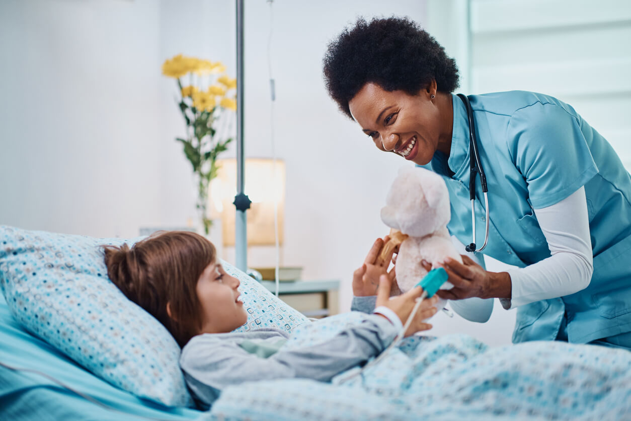 A black medical professional giving a teddy bear to a child patient in bed.