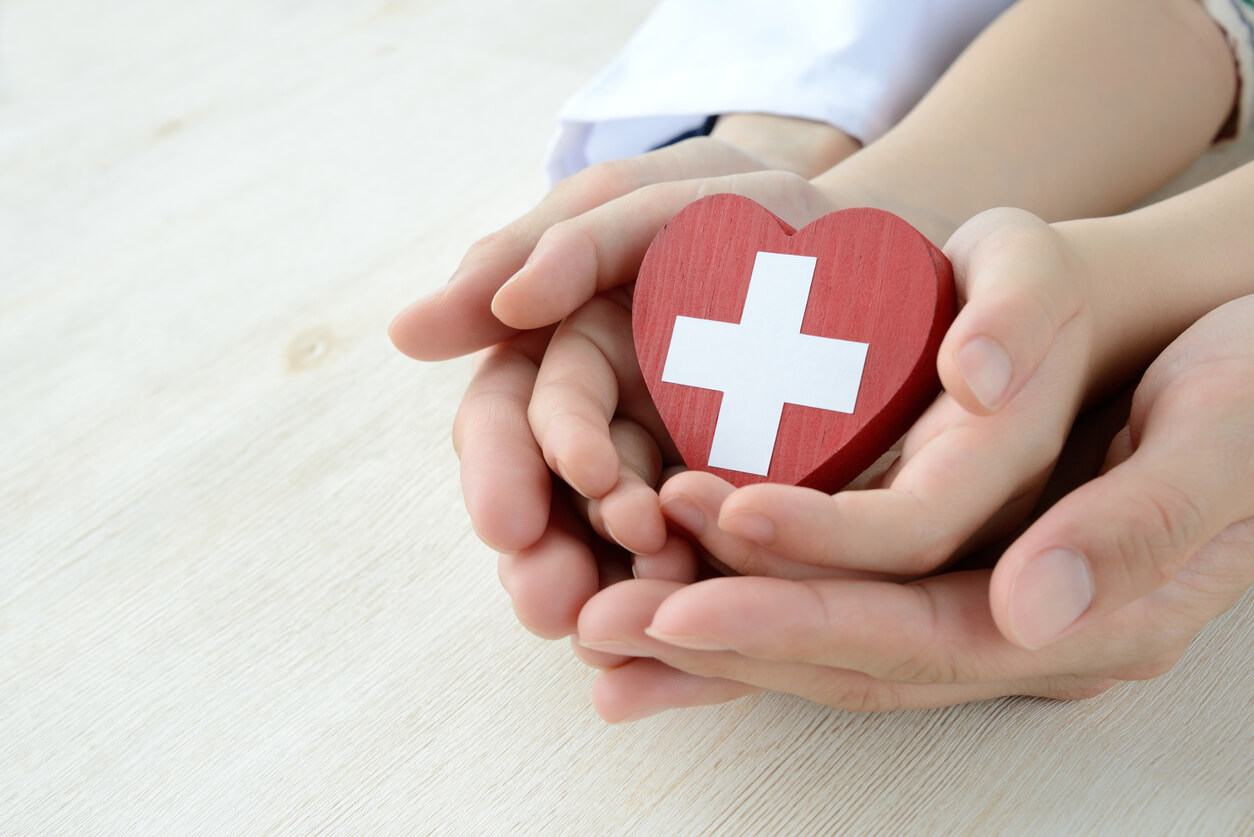 Händerna på en vuxen och ett barn som håller ett rött trähjärta med ett vitt kors i mitten.