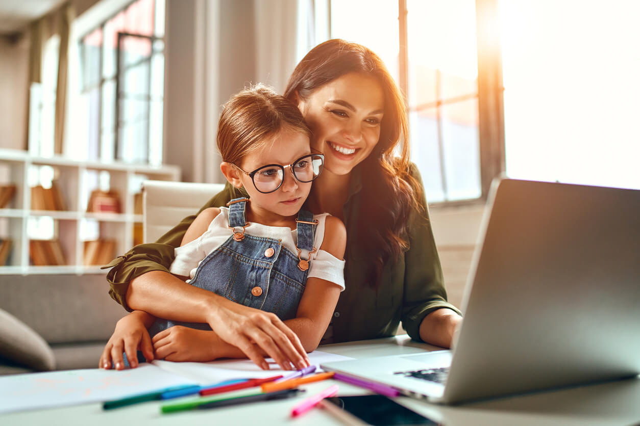 A mother studying online while her young daughter sits on her lap.
